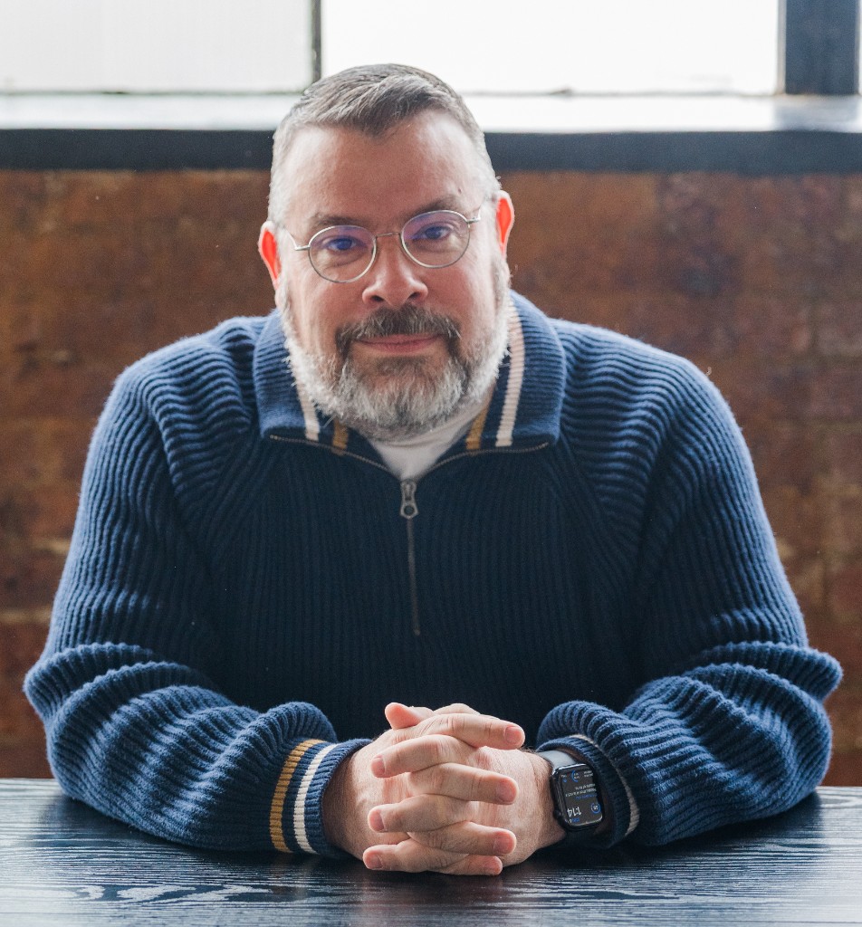 Tom Manners of Liquid Networks, seated at a table in a navy sweater, looking toward the camera.